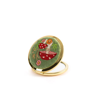 Opened round compact mirror with gold rim features a green velvet cover adorned with colorful mushroom and flower embroidery
