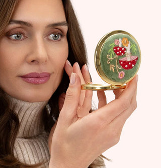 Model holds a round compact mirror with gold rim that features a green velvet cover adorned with colorful mushroom and flower embroidery