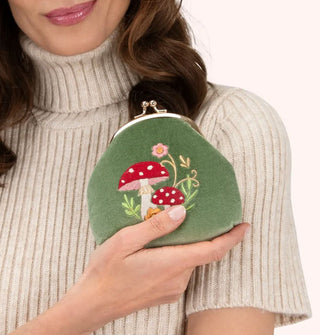 Model in beige ribbed turtleneck sweater holds a green velvet kiss lock change purse that features an embroidered design of two red and white spotted mushrooms among delicate ferns and flowers