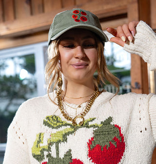 Model poses with hand raised to the brim of a sage green baseball cap with bright red tomato embroidery, worn with a white sweater also featuring tomato design