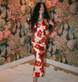 Model holding wine goblet wears a satin midi-length tomato print dress against a floral backdrop