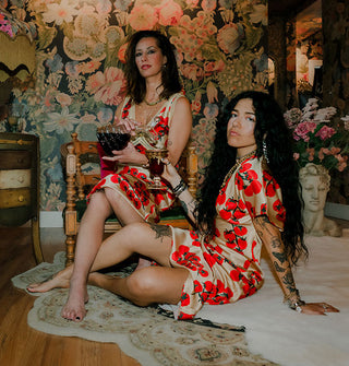 Two models wearing satin tomato print garments sit on a chair and rugged floor respectively against a floral backdrop staged with stone bust and side table