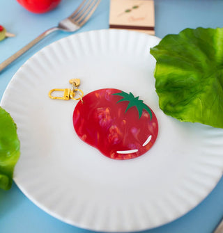 A mottled red plastic tomato keychain with green stem detail and gold clasp rests on a white paper place flanked by green lettuce leaves on a blue tabletop with a fork and matchbook in the background