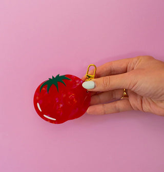 Model's hand holds a red and green tomato keychain with gold clasp against a pink backdrop