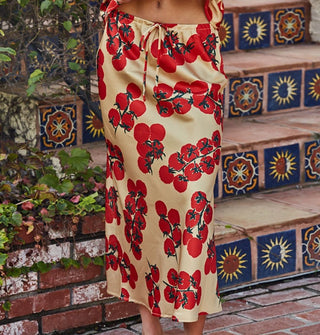 Model wears a tan satin midi-length skirt with tie front and print of red tomatoes on the vine against a Tuscan-style backdrop