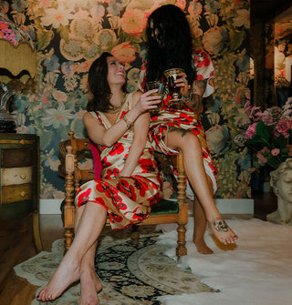 Two smiling models wearing satin tomato print garments and sharing a chair make a toast against a floral backdrop