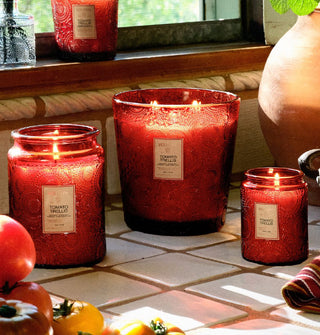 Four lit Tomato Trellis Voluspa candles, all in red embossed glass vessels, staged on a tiled countertop and windowsill with tomatoes, ceramic urn, and cloth towel