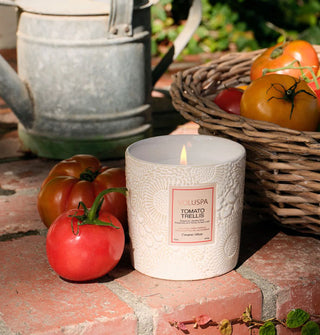 A lit Tomato Trellis Voluspa candle is staged on a brick surface alongside loose and basketed tomatoes and a metal watering can