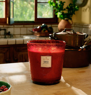 Lit Tomato Trellis Voluspa candle in a red embossed glass vessel is staged on a butcher block kitchen countertop with pots and pants and a bowl of cherry tomatoes with a tiled basin area under a window in the background