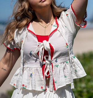 Model standing along the coast wears a white top with tie front, red trim, floral embroidery, and a peplum waist