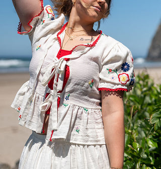 Model standing along the coast wears a white top with tie front, red trim, floral embroidery, and a peplum waist