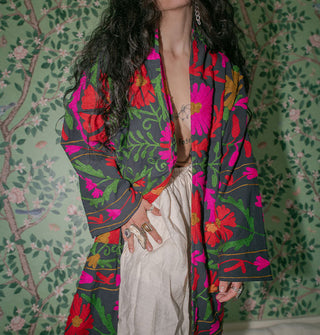 Model wears a long dark coat with brightly colored embroidery against a botanically wallpapered backdrop