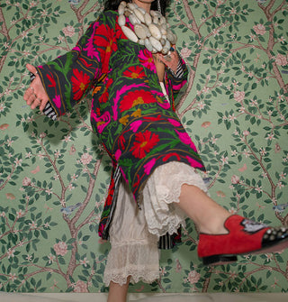Model kicking out foot with red loafer wears a long dark coat with brightly colored embroidery against a botanically wallpapered backdrop