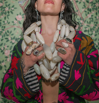 Model standing against a botanically wallpapered backdrop clutches an oversized wrapped beaded necklace she wears with a dark coat with brightly colored embroidery