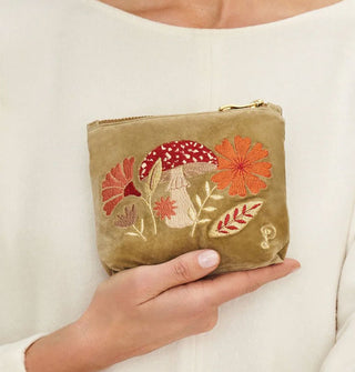 Model holds an olive green pouch that features embroidery depicting a red and white amanita mushroom flanked by flowers and leaves in an autumnal color palette