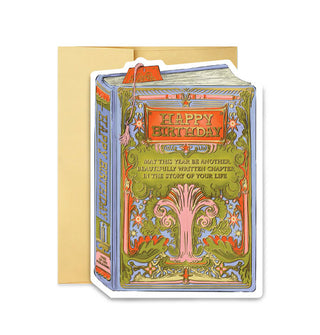 Greeting card in front of a gold envelope is designed to resemble a book cover that says, "Happy Birthday" in an orange rectangle with gold frame above a gold-accented tree that says, "May this year be another beautifully written chapter in the story of your life" in gold lettering; its spine says, "Happy Birthday" and "Tree of Life Publishing"