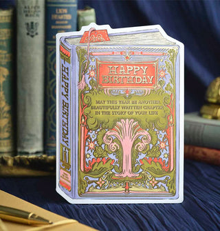 Greeting card is designed to resemble a book cover that says, "Happy Birthday" in an orange rectangle with gold frame above a gold-accented tree that says, "May this year be another beautifully written chapter in the story of your life" in gold lettering; its spine says, "Happy Birthday" and "Tree of Life Publishing"