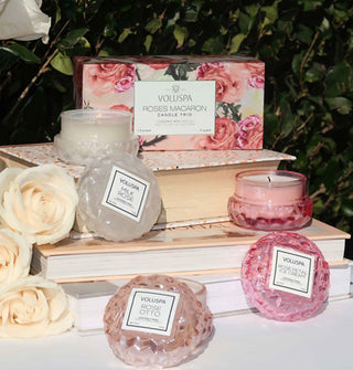 Lit Voluspa macaron candles in faceted glass jars are staged o a stack of books against a backdrop of greenery