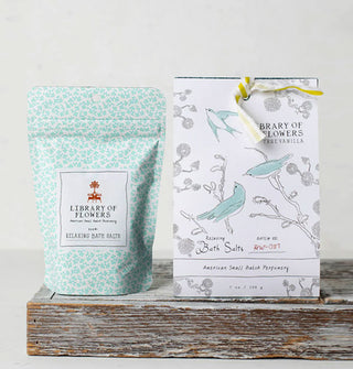 Inner and outer pouches of Library of Flowers True Vanilla Bath Salts featuring blue bird and flower motifs rest on a rustic wooden platform