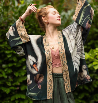 Model with arms extended upward wears a cropped kimono-style garment with a large swan design wrapped around it complemented by contrasting printed borders and flanked by flowers and a small cygnet