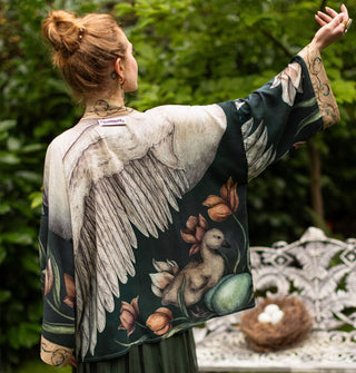 Model facing away from the camera and wearing a dark cropped robe-style top designed with a large swan and cygnet design outstretches one arm to show sleeve details