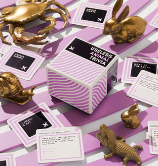 A box of Useless Animal Trivia rests on a ridged purple and white surface with sample cards and gold animal figurines