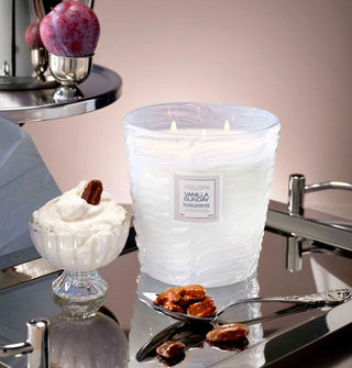 Lit three-wick Vanilla Sunday Voluspa candle on a reflective metal tray with candied nuts, a bowl of vice cream, a plum, and other items