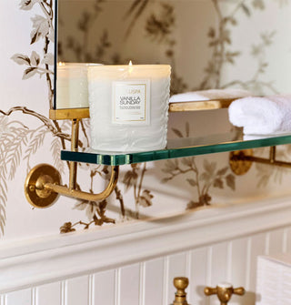 A lit single-wick Vanilla Sunday candle rests on a glass vanity shelf above a sink basin against a brown and white botanical wallpapered wall with white wainscoting below