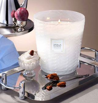 Lit Vanilla Sunday Voluspa candle in a giant frosted white embossed glass vessel rests on a metal tray with spoon, nuts, ice cream dish, and a plum and teapot nearby on a silver pedestal
