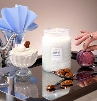 A lit single-wick Vanilla Sunday Voluspa candle in frosted white embossed glass jar rests on a metal tray with spoon, candied nuts, vanilla ice cream in a dish, fanned blue napkin, teapot, and model's hand reaching into the frame to place a plum on the tray