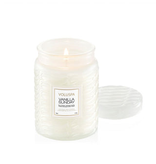 Lit single-wick Vanilla Sunday Voluspa candle in frosted white embossed glass jar with matching lid set to the side