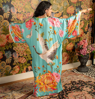 Model with back to camera and arms outstretched wears a long, flowing blue robe with bold floral print around a large crane with wings spread