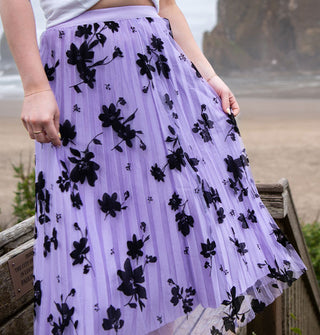 Model wears a pleated purple tulle skirt with black floral embroidery on a beach boardwalk