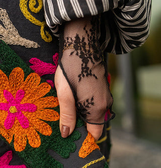 Closeup of the sleeve with thumbhole of a sheer black floral lace top layered with a colorfully embroidered coat with striped cuff
