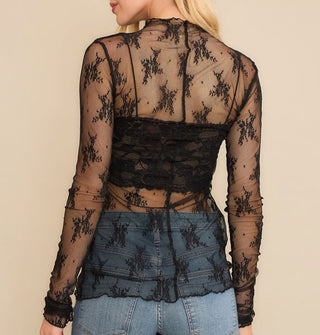 Model facing away from the camera wears a long-sleeved sheer black floral lace top with black bralette and blue jeans