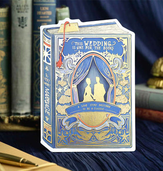 Blue greeting card features a gold silhouette of a couple inside an ornate gold-accented, curtained cameo frame over which a banner reads, "A love story destined to be a classic," and the words, "This wedding is one for the books" at the top; on the spine is the message, "A very merry marriage" in gold
