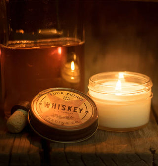 A lit Whiskey jar candle with lid set to the side is staged with a clear flask that reflects the flame