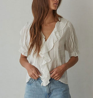Model wears a ruffly white blouse with pleated details and short balloon sleeves