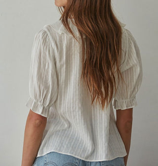 Model with back to camera wears a ruffly white blouse with a vertical weave pattern and short balloon sleeves