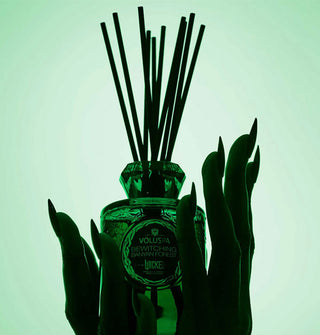 Two hands with long fingernails cradle a green glass Voluspa oil diffuser bottle with black reeds inserted
