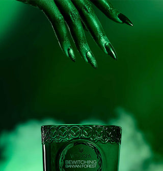 A green hand with long fingernails extends down toward a green glass Bewitching Banyan Forest candle set against a smoky backdrop