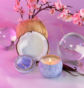 Small lit purple Wicked edition Voluspa candle tin with adjacent lid is staged with glass orbs, a halved coconut, vanilla stalks, and pink cherry blossoms on a purplish-pink backdrop