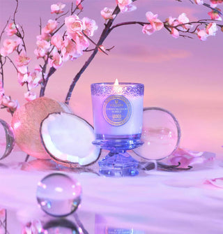 Lit Cherry Blossom Bubble Voluspa candle  atop pedestal lid is staged with glass orbs, halved coconut, and pink cherry blossom branches on a misty surface against a pink backdrop