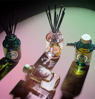 Pink and green glass reed diffuser bottles on a heavily shadowed pink and green surface, two with reeds inserted