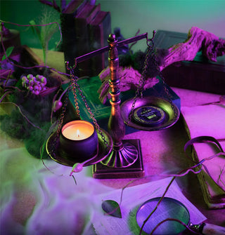 Lit Wicked edition Voluspa Palo Santo Spells candle tin and its lid rest on either side of an old-fashioned scale surrounded by objects of intrigue in rippling mist and green and purple light