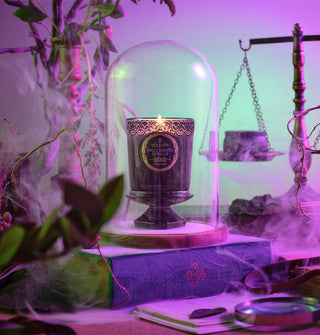 A lit Wicked edition Palo Santo Spells Voluspa candle jar is encased in a glass cloche and staged with objects of intrigue among swirling mist or smoke