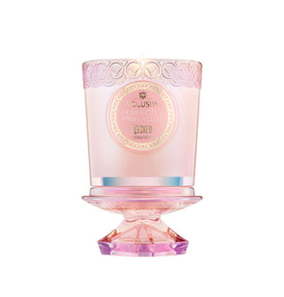 Lit pink glass Voluspa candle jar with decorative label is placed atop its pedestal-shaped lid