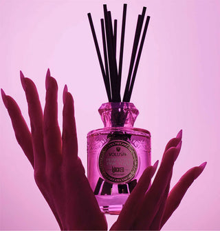 Two hands with long fingernails cradle a pink glass Voluspa oil diffuser bottle with black reeds inserted