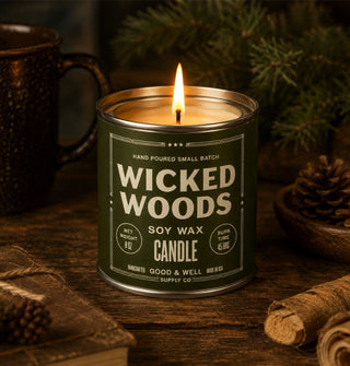 Lit Wicked Woods Soy Wax paint can-style candle is staged on a rustic wooden surface with pine branch, pine cone, coffee mug, antique book, and other items