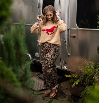 Model standing in front of an Airstream trailer wears a beige sweater with galloping red horse design paired with leopard print pants and copper clogs